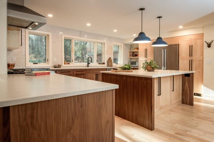 Angled view of a modern kitchen remodel in Hollis, NH by Blackdog Design/Build/Remodel, highlighting an oversized island with a white quartz countertop, walnut cabinetry, pendant lights, and abundant natural light.