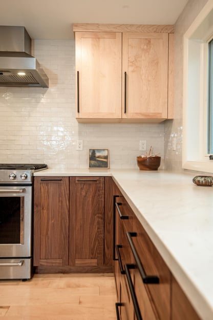 Close-up of a modern kitchen remodel in Hollis, NH by Blackdog Design/Build/Remodel with walnut base cabinets, light maple upper cabinets, white quartz countertops, white tile backsplash, and stainless steel range.