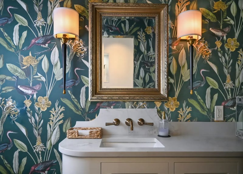 Elegant bathroom featuring a vanity with a marble sink, brass fixtures, green botanical and bird-themed wallpaper, and an antique-style mirror flanked by two wall sconces
