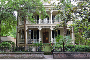 Historic two-story plantation-style home with upper and lower covered decks and large columns. Large trees on either side and gate in front of house.