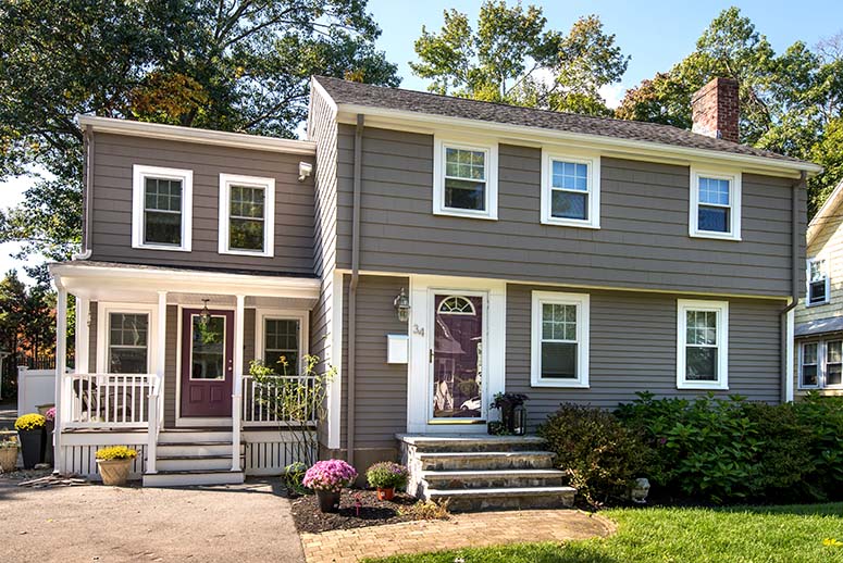 Remodeled home exterior with addition on the left side. Two-story brown/gray home with white trim and small front porch on the addition. Trees in background.