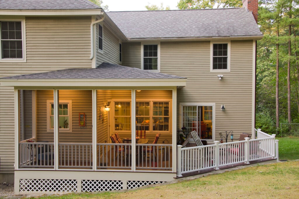 Screened-in porch added to the back of a large two-story beige home, with open deck to the right side, lights on in the porch, trees behind home.