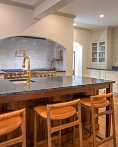 New kitchen island with woodgrain cabinets, black/gray soapstone countertop, stool seating along one side. View of large gas range and oven on far side behind the island. Wood floors.