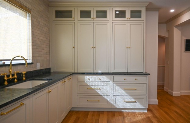 Remodeled kitchen with white cabinets, black/gray soapstone countertop, gold faucet and fixtures, wood floors, white brick backsplash and window behind counter and sink.