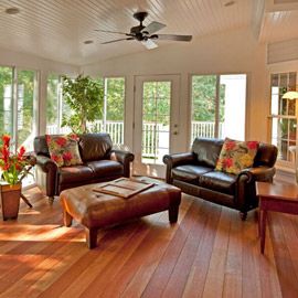 home addition living space with wood floors, two leather couches and an ottoman, white wood beam ceiling and wall of windows in background with door leading out to a deck.