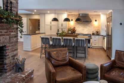 A newly-trasformed open-concept living room and kitchen remodel in Andover, MA looking beyond the fireplace and onto the kitchen's large center island with countertop seating, black pendant lighting, white cabinets, and stainless steel appliances