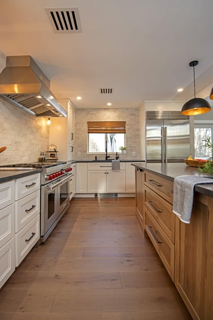 A view toward the sink underneath a window beyond the large center island and stainless steel range among wide-plank wood flooring in a kitchen remodel in Andover, MA by Blackdog Design/Build/Remodel