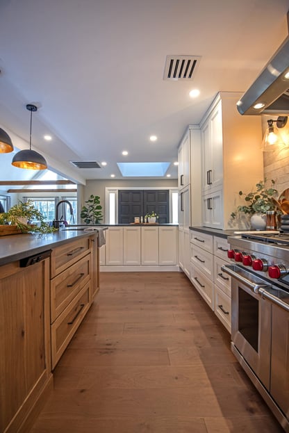 A view from a remodeled kitchen in Andover, MA looking toward the front door of the home featuring wide-plank wood flooring, white cabinetry, a large center island illuminated by black pendant lighting, and a stainless steel range