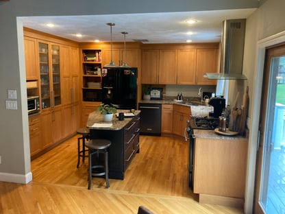 A "before" perspective preceding a remodel transformation by Blackdog Design/Build/Remodel showing a dated kitchen with honey-toned wood cabinets, granite countertops, a small center island, and mixed appliances opening into an adjacent living area with hardwood floors.