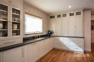 A kitchen remodel project in Andover, MA featuring white shaker cabinets, brass knobs and pulls, and dark stone countertops. A farmhouse-style sink sits beneath a window with a woven shade and white subway tile backsplash.