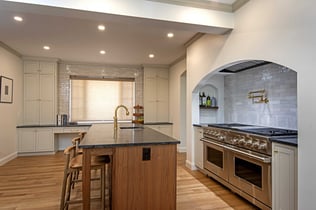 A large center island with counter seating sits in front of a stainless steel range underneath a custom archway in a remodeled kitchen in Andover, MA by Blackdog Design/Build/Remodel