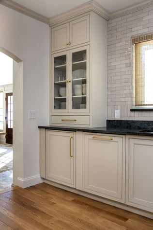 Corner view of a kitchen remodel in Andover, MA featuring shaker-style cabinets with glass upper cabinets holding dinnerware beside an archway opening up to the home's entryway.