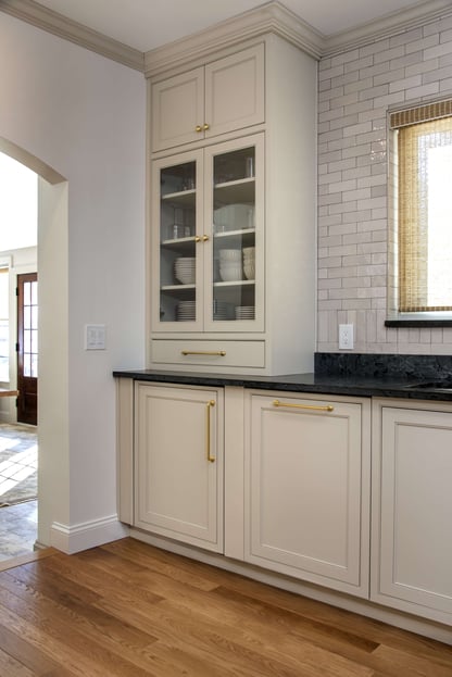 Corner view of a kitchen remodel in Andover, MA featuring shaker-style cabinets with glass upper cabinets holding dinnerware beside an archway opening up to the home's entryway.