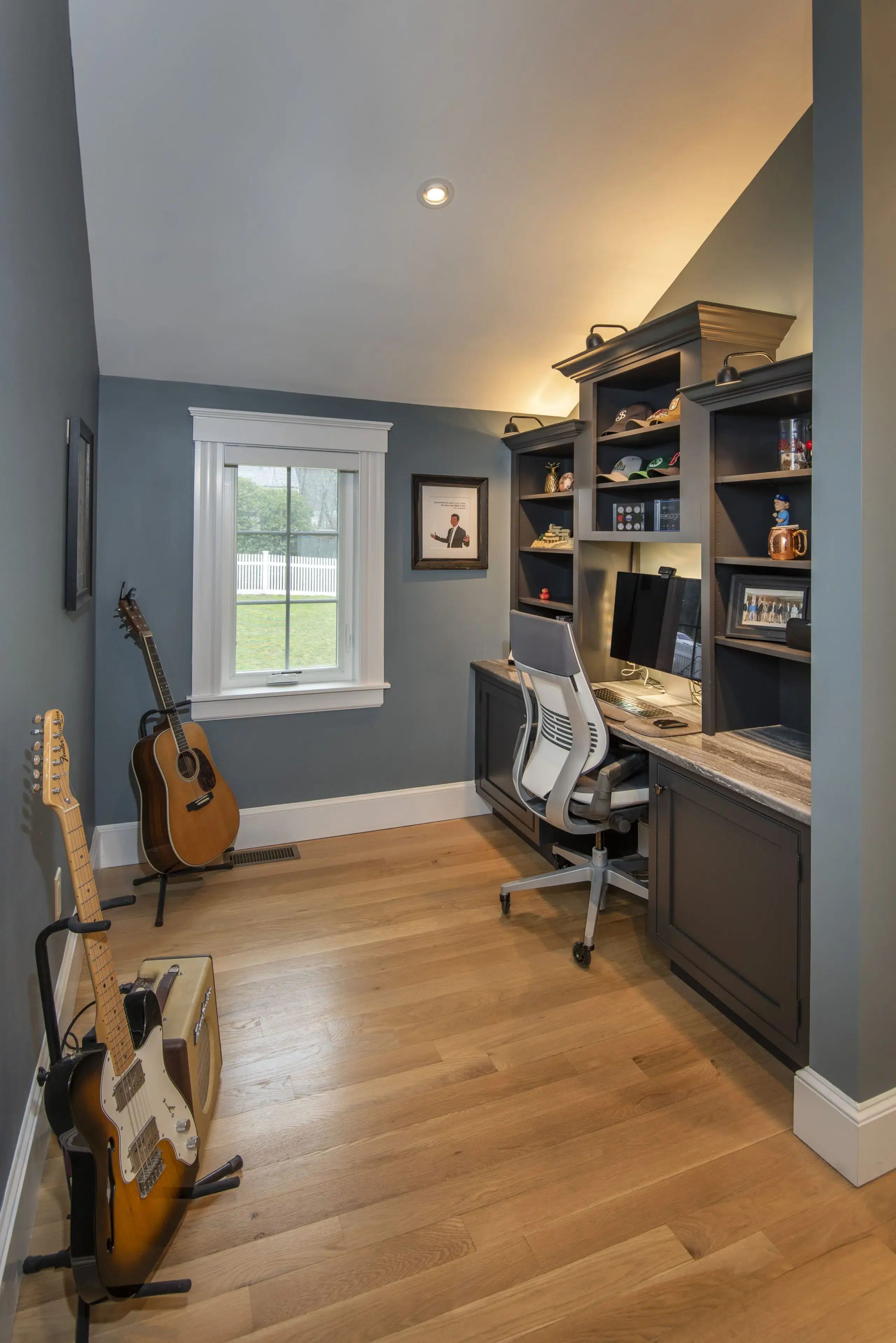 Home office addition with wood floors, built-in desk along right side with desk chair pushed in, facing built-in shelves on wall above desk. Small double-hung window on far wall. Two guitars on stands against left-side wall.
