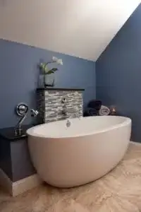 Remodeled bathroom with blue walls, tan tile flooring, separate white soaking tub with showerhead attachment on wall above.