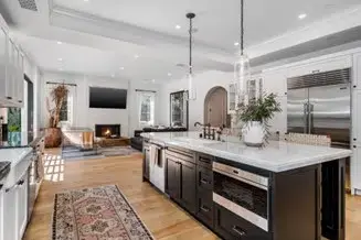 Open-concept kitchen and living room remodel with long island in foreground with black cabinets, white stone countertop. Wood floors. High ceilings. Recessed lighting.