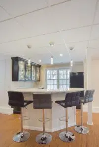 Finished basement area with wood floors ,white walls, and small bar area with white countertop, black stools around island, and black cabinets against far wall.