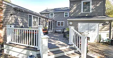 Back exterior view of a two-story gray home with raised wood deck with white railings and few steps in foreground leading to a patio area.