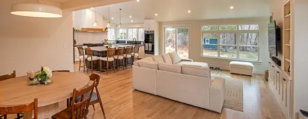 open concept floor plan remodel with wood floors, open dining, living, kitchen areas with recessed lighting, white sectional couch, and row of double-hung windows along far wall.