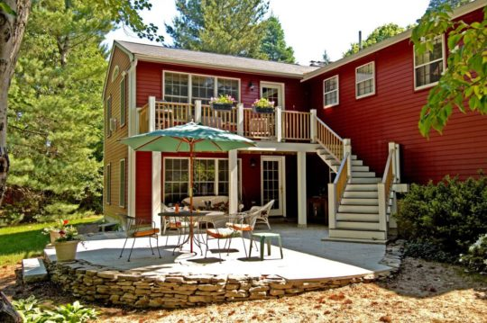 Backyard patio with chairs, table & umbrella at a red, two-story home with stairs leading to upper deck. Trees surrounding.