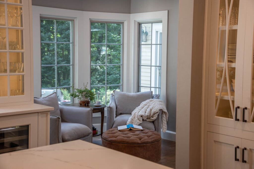 Cozy reading corner in a remodeled home. Tall corner windows with view of trees outside. Two cushioned gray chairs in front of windows, facing round cushioned ottoman.