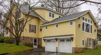 A large historic home with A-frame roof, yellow siding, dark shutters, and stone foundation. Two-car garage with white doors.