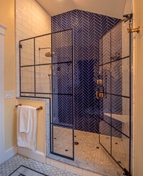 Glass-enclosed walk-in shower with blue grids on glass door and herringbone style dark blue tile on one shower wall, as accent.