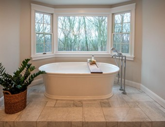 A white free-standing tub on a light gray tile floor, under a white bay window with view of trees outside.