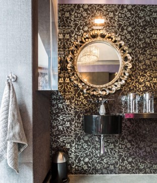 A small bathroom vanity with bold black-and-white patterned wallpaper on the wall, with a round silver-framed mirror above the sink.