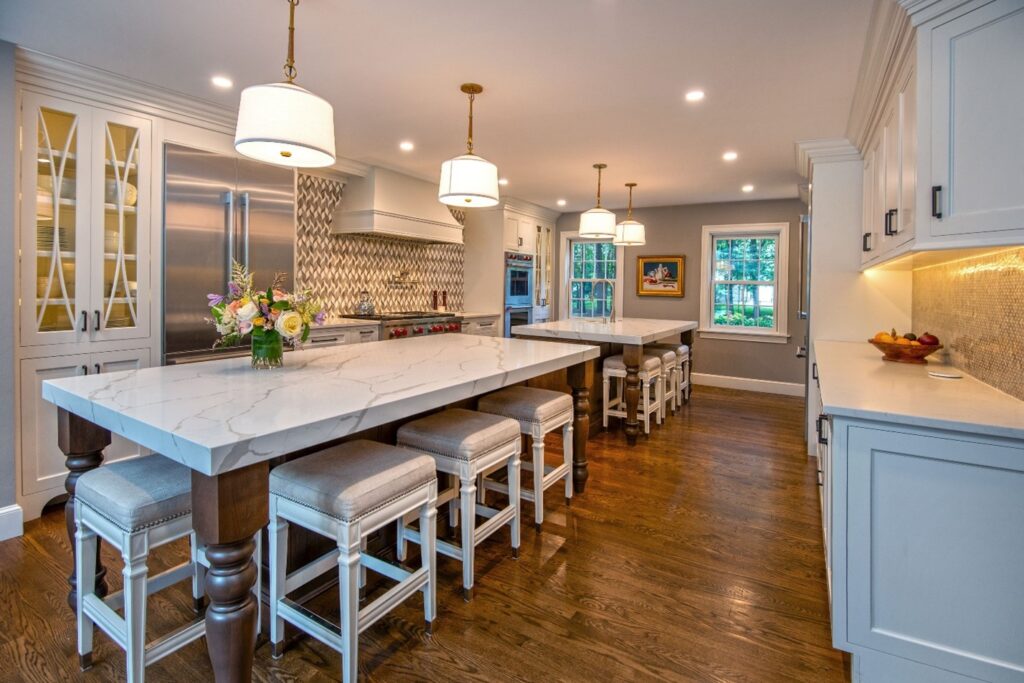 Remodeled kitchen with white cabinets, two long islands down the middle with white slab marble countertops, wood floors, cushioned stool seating at both islands, pendant lights and recessed lighting.