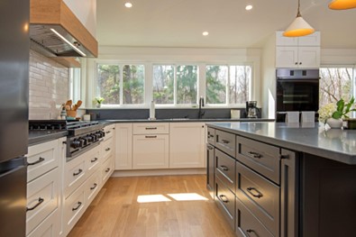 Remodeled kitchen with white cabinets along wall, gray cabinets for island, gray stone countertops, light wood floors, and row of white-framed sliding windows above counter on far wall. Recessed lighting.